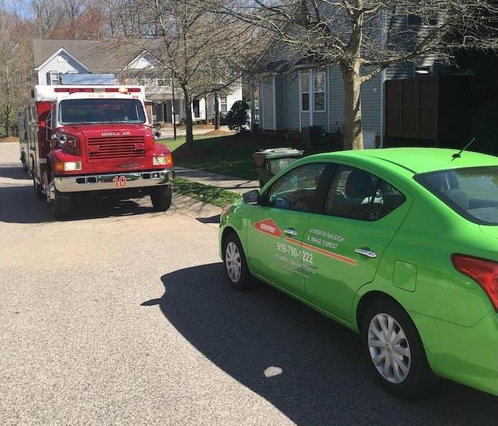 green SERVPRO car and red emergency vehicle sitting on the road in front of a home