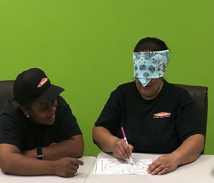 Two people sitting at a table, one is blindfolded and holding a pen to a paper while the other instructs what to draw
